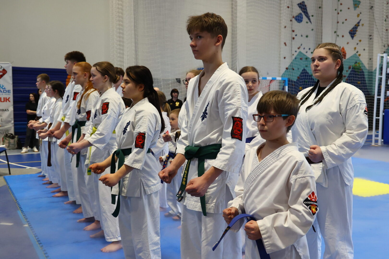 Group of young martial artists in white gi with green belts lined up on blue mats, indoors for a ceremony or training session.