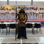 Samurai warrior in ornate armor stands with a polearm in front of a line of karate students on a competition mat stage.