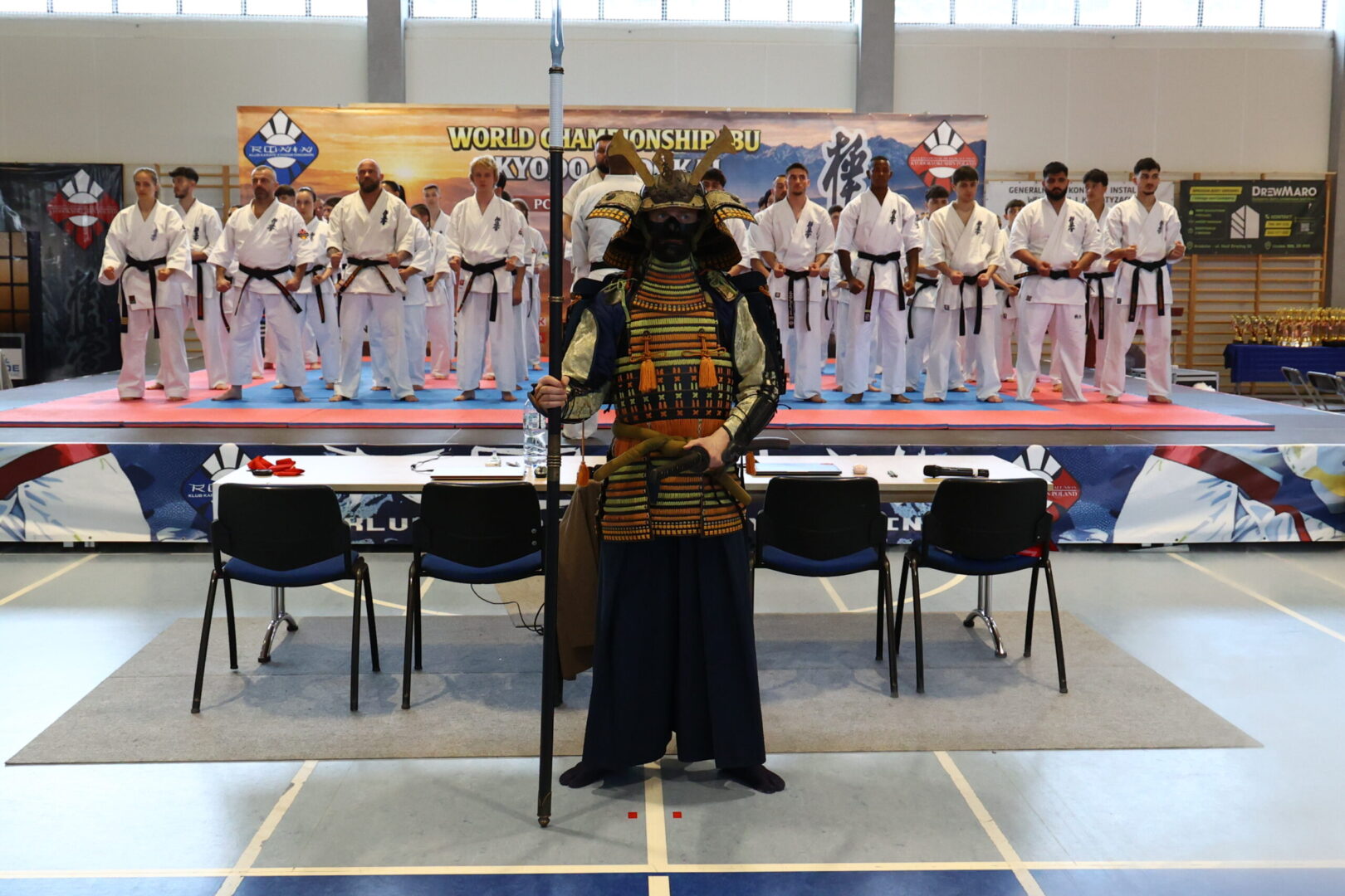 Samurai warrior in ornate armor stands with a polearm in front of a line of karate students on a competition mat stage.