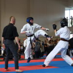Two karate competitors in white uniforms kick at each other on blue and red mats as a referee watches nearby.