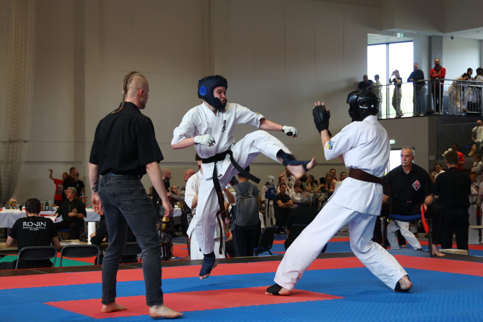 Two karate competitors in white uniforms kick at each other on blue and red mats as a referee watches nearby.