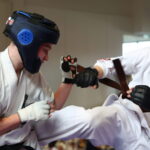 Two martial artists in white gis sparring; one is delivering a high kick while wearing blue headgear and gloves.