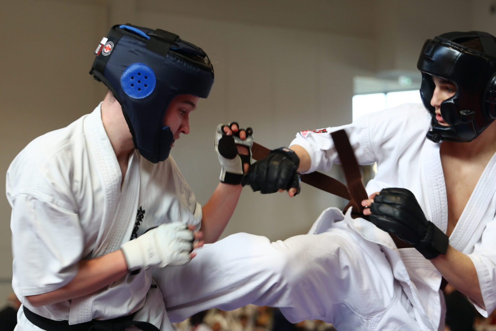 Two martial artists in white gis sparring; one is delivering a high kick while wearing blue headgear and gloves.