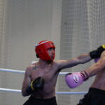 Boxers wearing headgear exchange punches in a ring; red-helmeted fighter lands a straight left on blue-helmeted opponent while a net backdrop is visible in the gym.