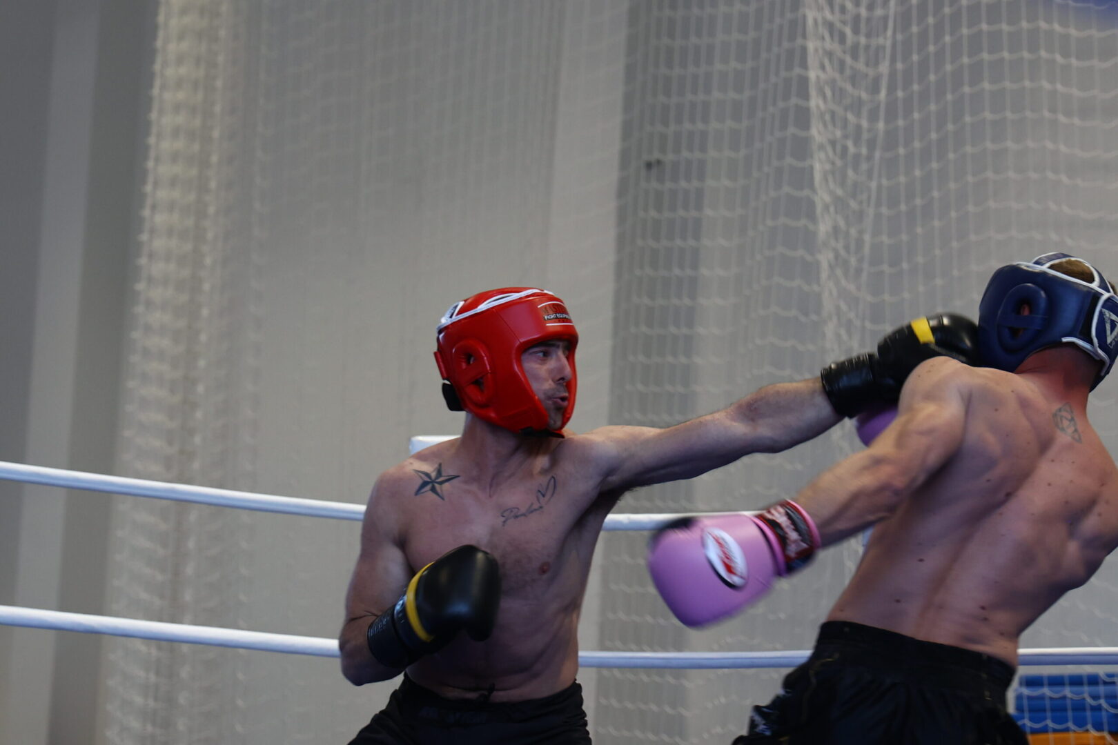 Boxers wearing headgear exchange punches in a ring; red-helmeted fighter lands a straight left on blue-helmeted opponent while a net backdrop is visible in the gym.