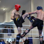 Two shirtless fighters in protective headgear trade kicks in a boxing ring as an official watches nearby.