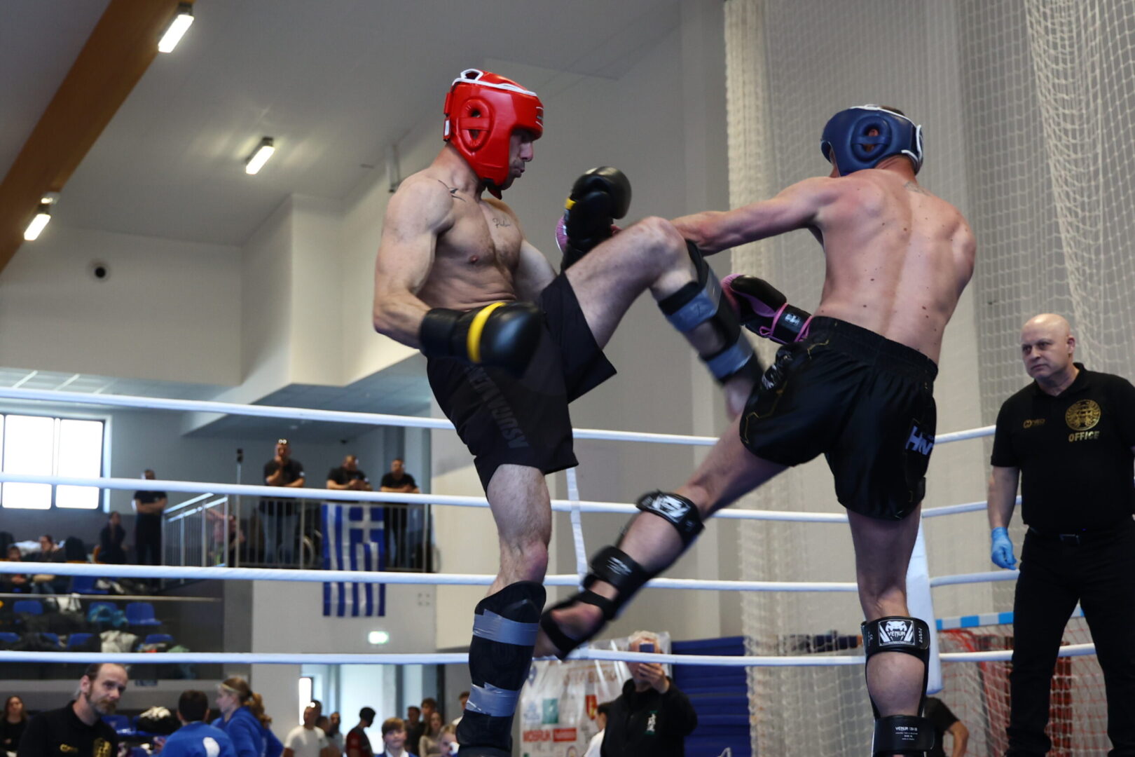 Two shirtless fighters in protective headgear trade kicks in a boxing ring as an official watches nearby.