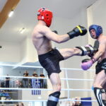 Two male fighters spar in a boxing ring; one with a red helmet delivers a high kick to a blue-helmeted opponent while spectators watch in the background.