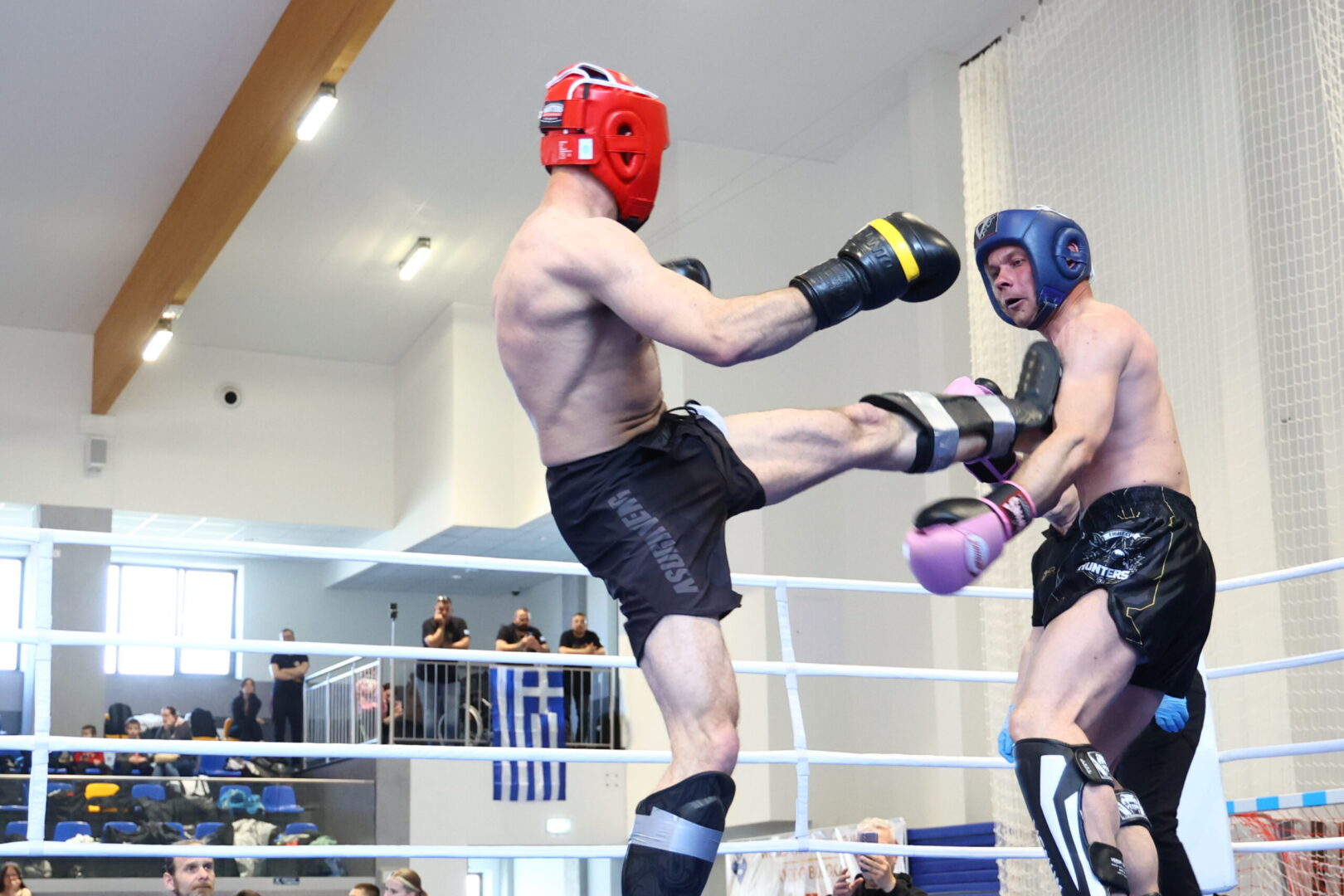 Two male fighters spar in a boxing ring; one with a red helmet delivers a high kick to a blue-helmeted opponent while spectators watch in the background.