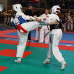 Two taekwondo practitioners in white uniforms and protective gear exchange a kick on multi-colored mats, with spectators watching from the stands in the background.