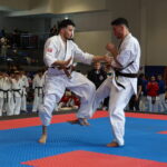 Two male karate practitioners spar on a blue-and-red tatami, one delivering a kick as the other blocks, with spectators and teammates in the background.