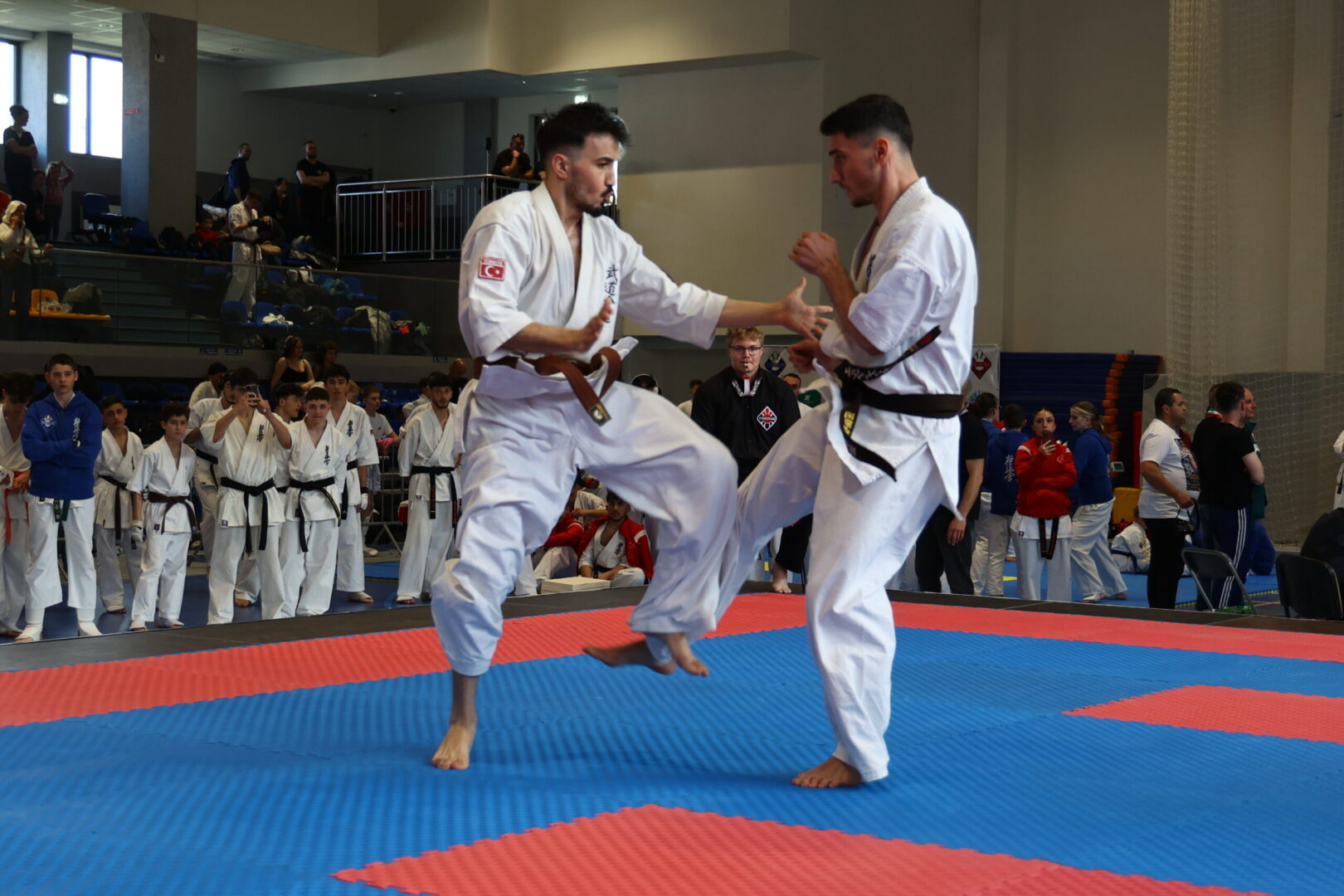 Two male karate practitioners spar on a blue-and-red tatami, one delivering a kick as the other blocks, with spectators and teammates in the background.