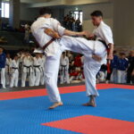 Two male judoka in white uniforms grapple on a blue-and-red tatami while spectators watch from the background.