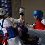 Two taekwondo athletes sparring in white uniforms with blue and red protective gear as one delivers a high kick toward the other in a gym.