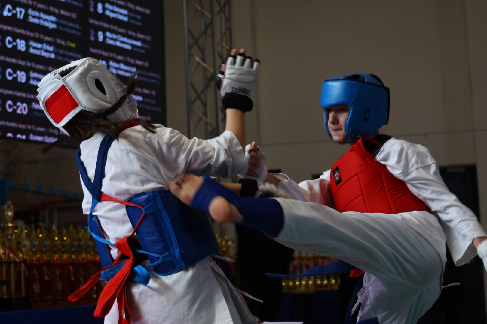 Two taekwondo athletes sparring in white uniforms with blue and red protective gear as one delivers a high kick toward the other in a gym.