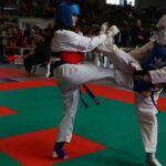 Two young taekwondo practitioners spar on a colorful mat, wearing protective headgear and chest guards in a crowded gym.
