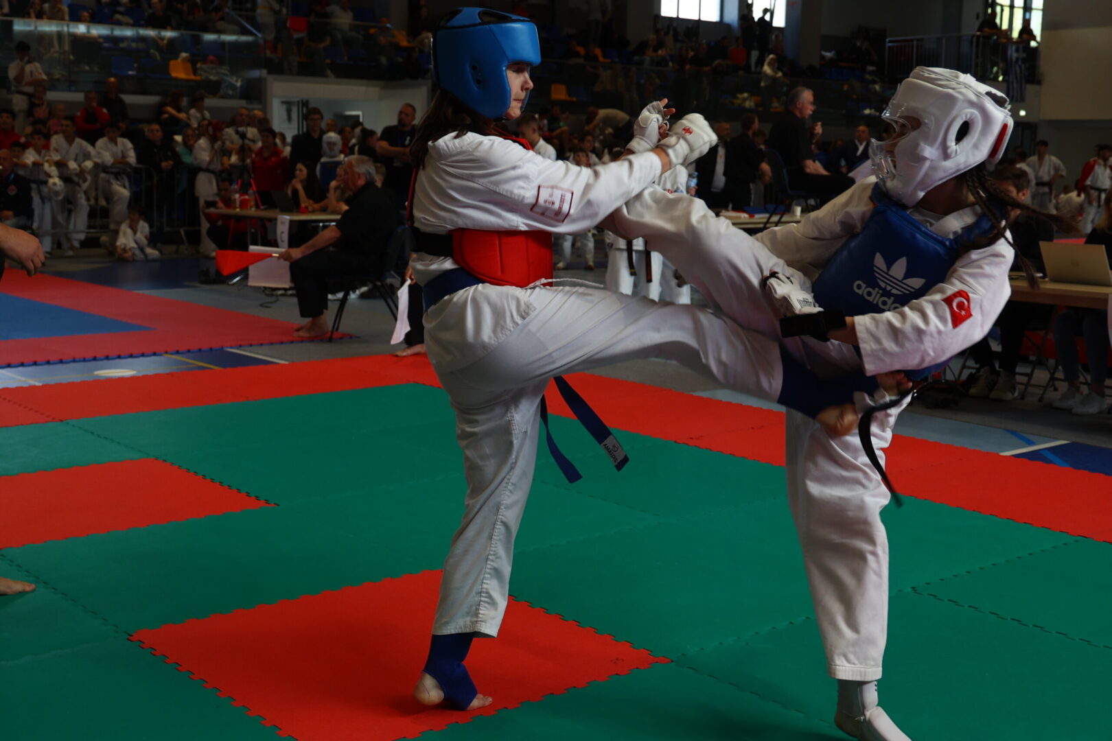 Two young taekwondo practitioners spar on a colorful mat, wearing protective headgear and chest guards in a crowded gym.