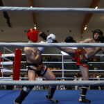 Muay Thai match in a boxing ring with two shirtless fighters; red headgear on the left fighter delivering a high kick to the right fighter in black helmet in red shorts.