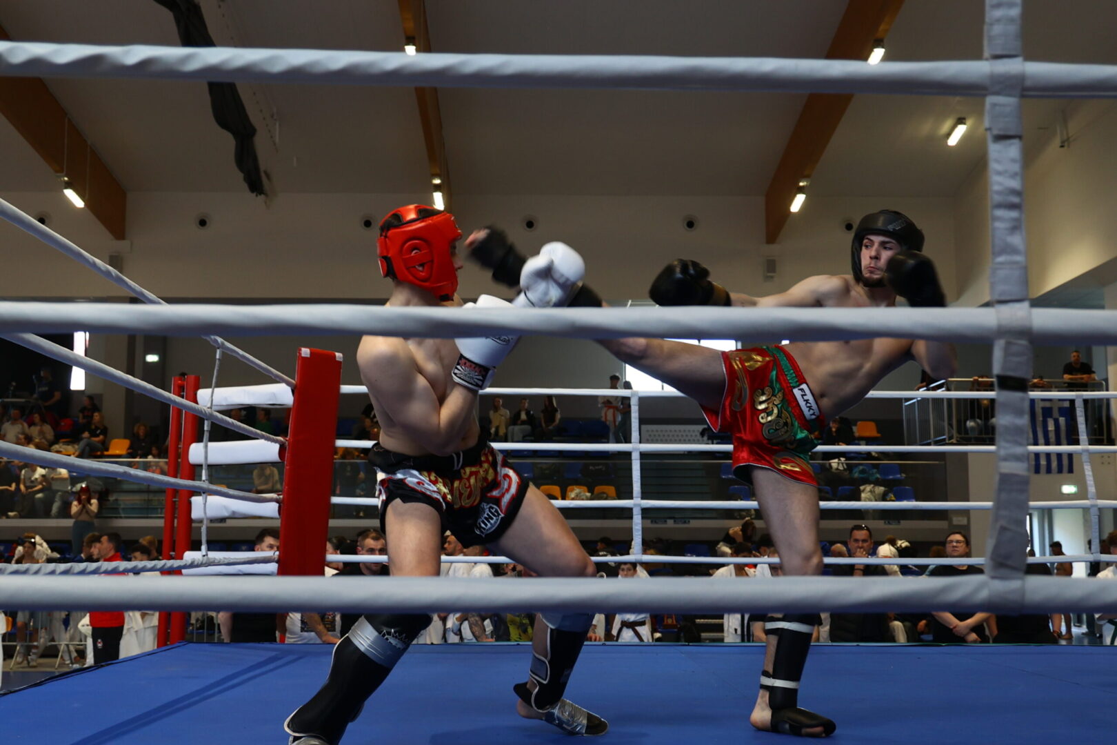Muay Thai match in a boxing ring with two shirtless fighters; red headgear on the left fighter delivering a high kick to the right fighter in black helmet in red shorts.