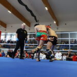 Two fighters grapple inside a boxing ring as the referee watches attentively. The boxer in green and red trunks clashes with a opponent in black trunks with red gear nearby a blue mat arena. A crowd sits and stands around the ring in a gymnasium.