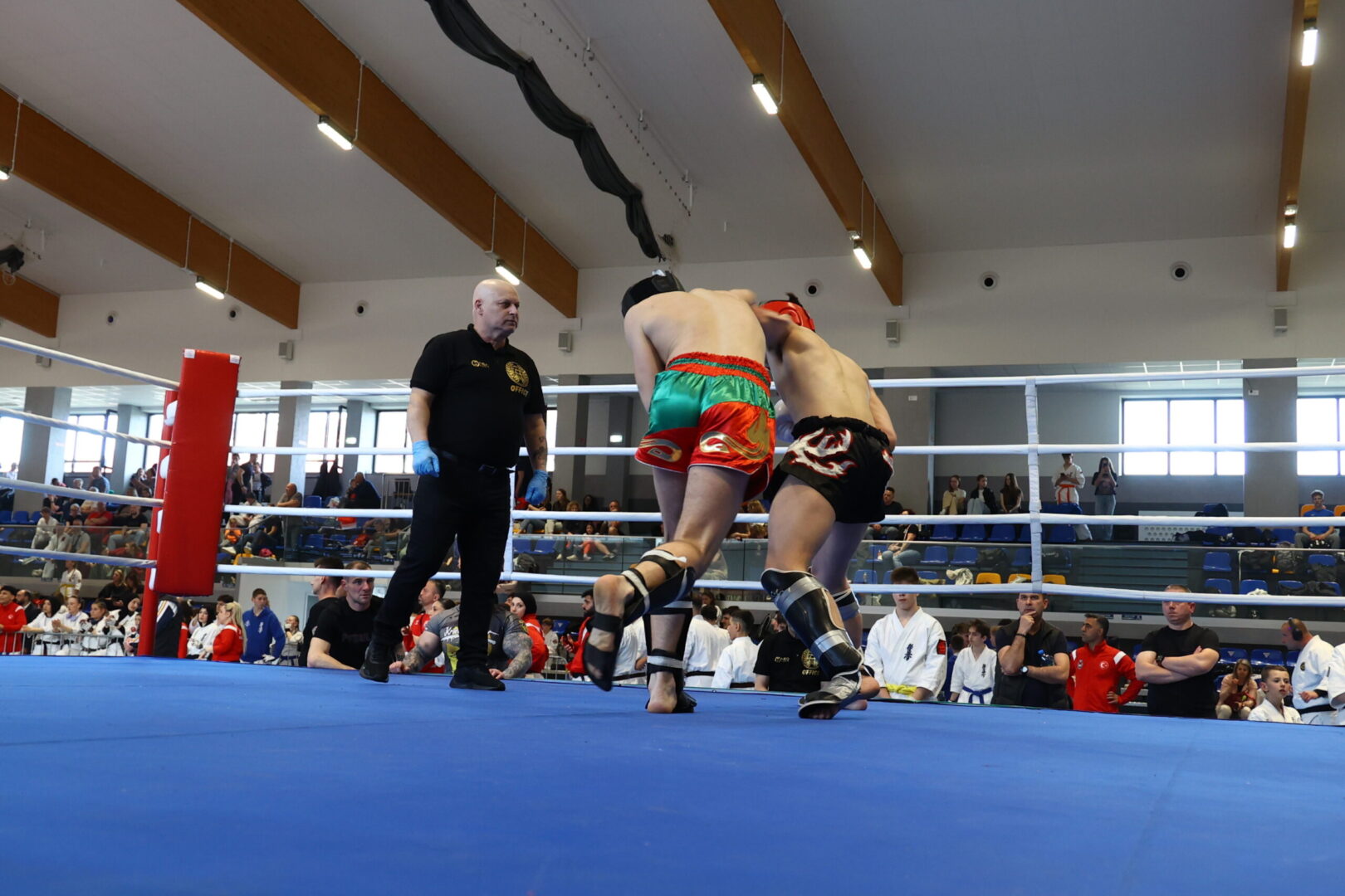 Two fighters grapple inside a boxing ring as the referee watches attentively. The boxer in green and red trunks clashes with a opponent in black trunks with red gear nearby a blue mat arena. A crowd sits and stands around the ring in a gymnasium.