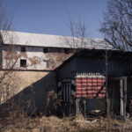 Abandoned industrial buildings with peeling plaster, a metal roof, and leafless trees against a clear blue sky.Conveys a derelict, neglected scene.