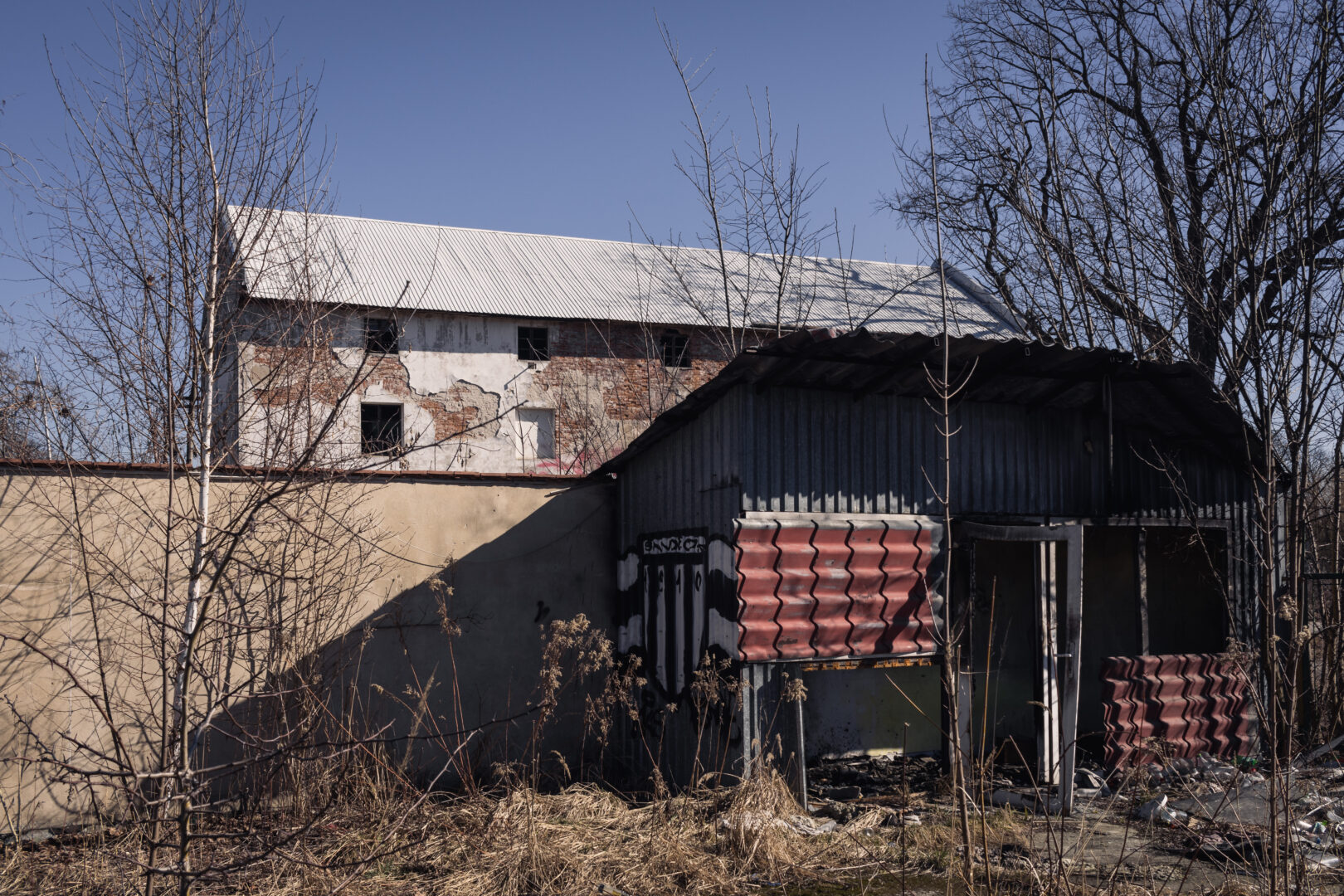 Abandoned industrial buildings with peeling plaster, a metal roof, and leafless trees against a clear blue sky.Conveys a derelict, neglected scene.