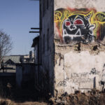 Graffiti-covered, crumbling building with a bold black-and-white monster mural; rubble at the base and a person walking on a distant walkway.