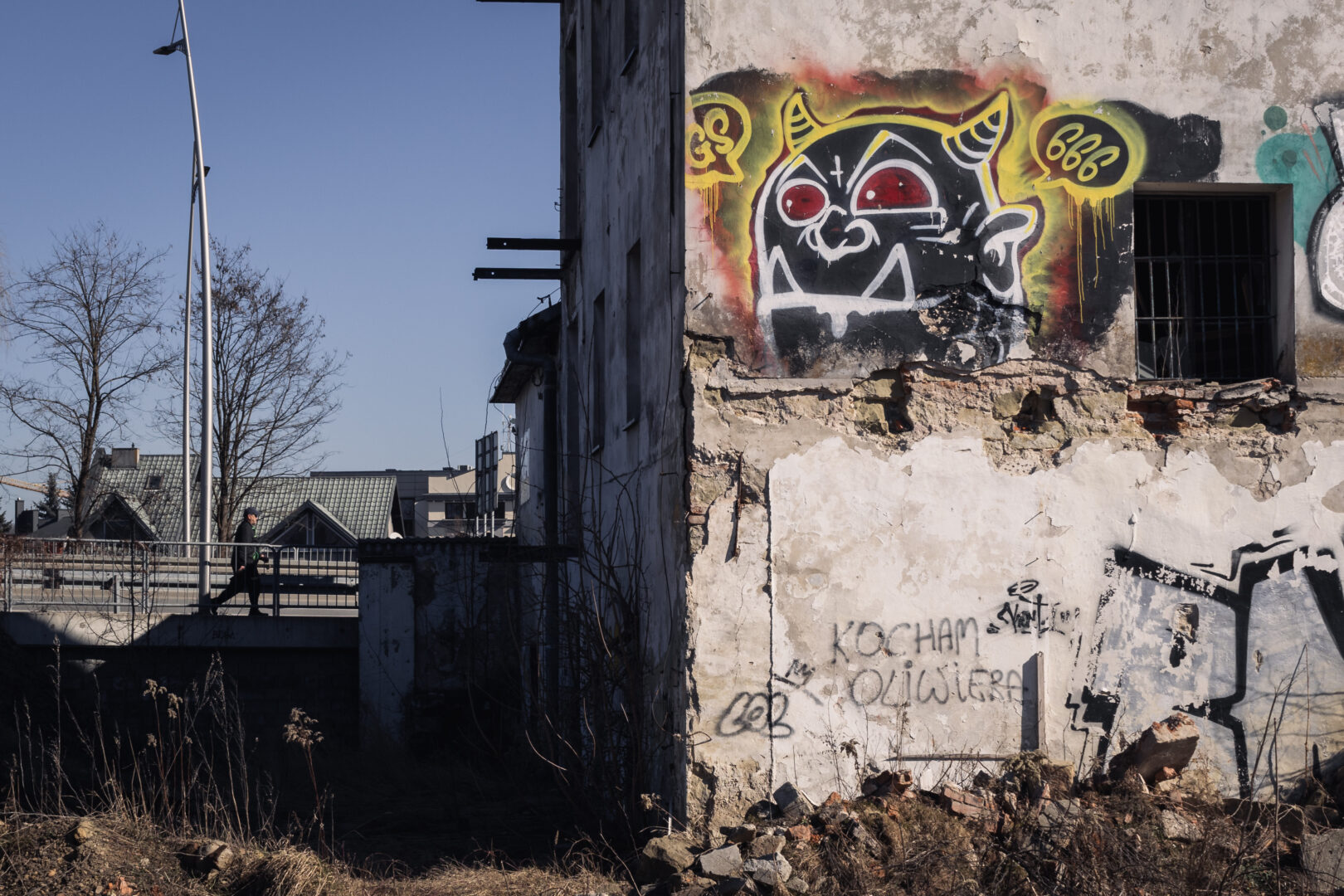 Graffiti-covered, crumbling building with a bold black-and-white monster mural; rubble at the base and a person walking on a distant walkway.