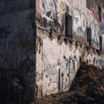 Crumbling concrete building wall with colorful graffiti, broken windows, and rubble on the ground in a neglected yard.
