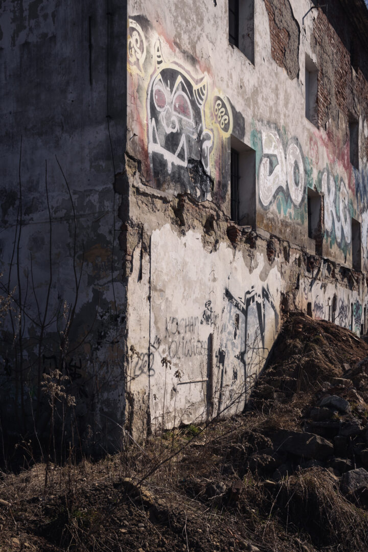 Crumbling concrete building wall with colorful graffiti, broken windows, and rubble on the ground in a neglected yard.
