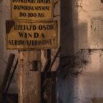 Two yellow, weathered warning signs in Polish mounted on a rough wooden post inside a dim, old workshop or warehouse.