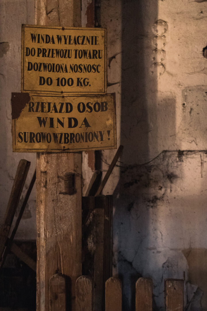 Two yellow, weathered warning signs in Polish mounted on a rough wooden post inside a dim, old workshop or warehouse.