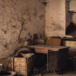 Dusty, abandoned workshop corner with cracked walls, wooden table, cabinet, and scattered debris including a tire and crates.