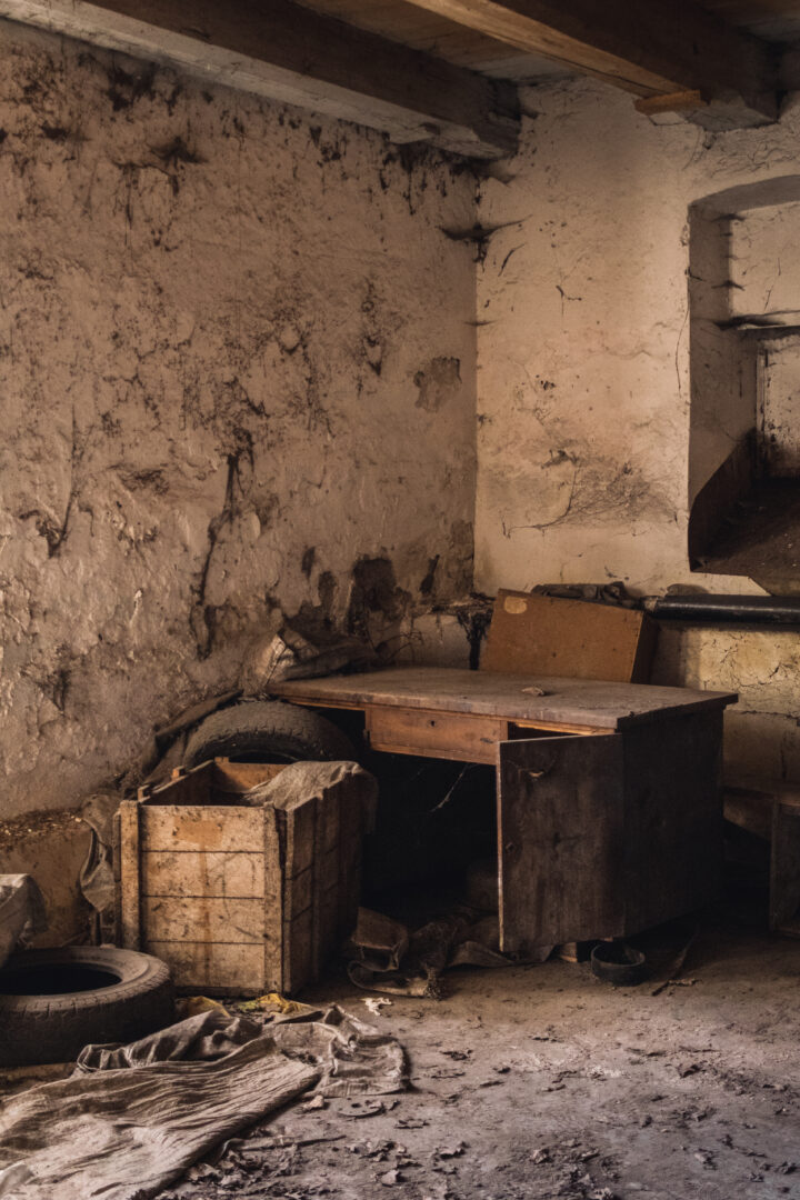 Dusty, abandoned workshop corner with cracked walls, wooden table, cabinet, and scattered debris including a tire and crates.