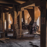 Abandoned factory interior with wooden support beams, dusty floor, and old machinery in a dim, sunlit room.