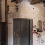 Dilapidated interior with a peeling gray metal door, graffiti on the walls, and rubble on the floor.