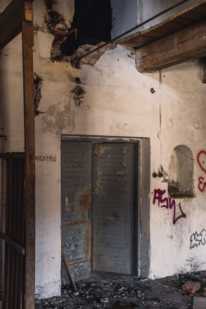 Dilapidated interior with a peeling gray metal door, graffiti on the walls, and rubble on the floor.