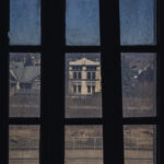 View through a dark window with three vertical panes, revealing a pale, colonial-style building across the street and bare trees in the foreground.