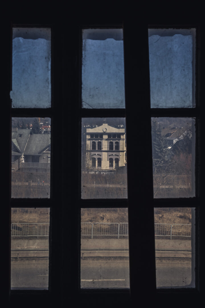 View through a dark window with three vertical panes, revealing a pale, colonial-style building across the street and bare trees in the foreground.