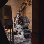 Industrial concrete mixer in a dusty, dim workshop with exposed wooden beams and caution tape surrounding the equipment.