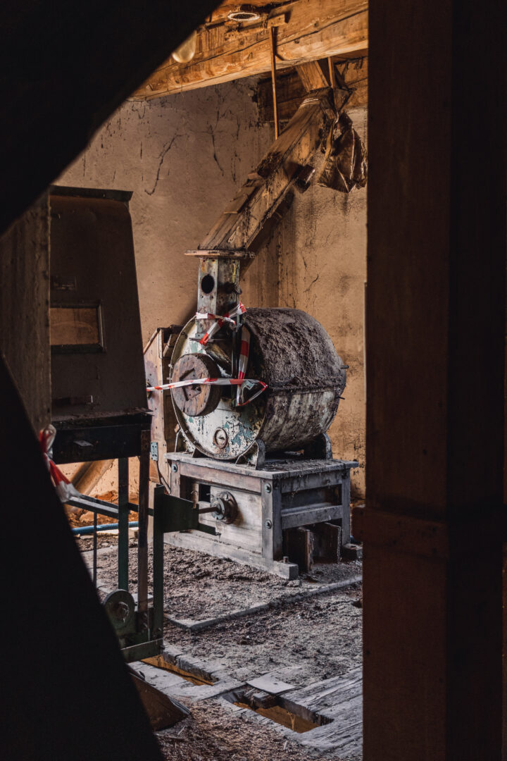 Industrial concrete mixer in a dusty, dim workshop with exposed wooden beams and caution tape surrounding the equipment.