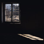 Dim interior with a single window letting in light; outside are bare trees and a white building, sunlight casts a patch on the floor.