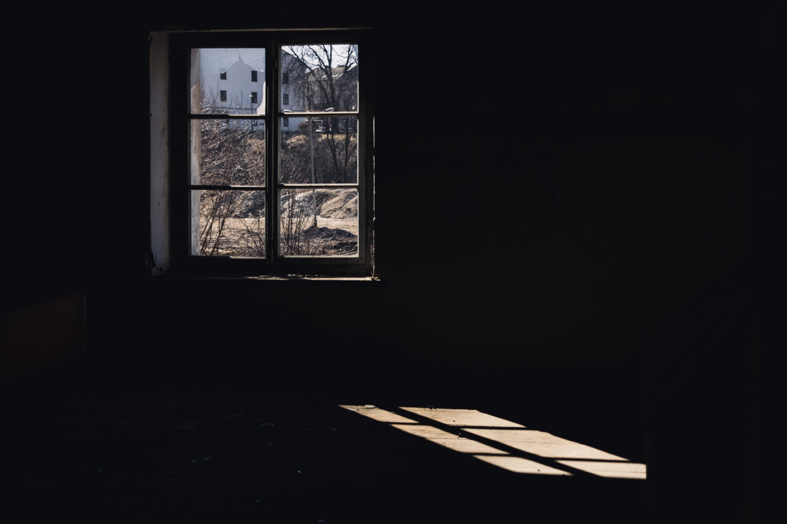 Dim interior with a single window letting in light; outside are bare trees and a white building, sunlight casts a patch on the floor.