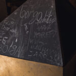 Dark, triangular metal panel covered in chalk graffiti with random words and markings; yellow base at the bottom, in a dim workshop.