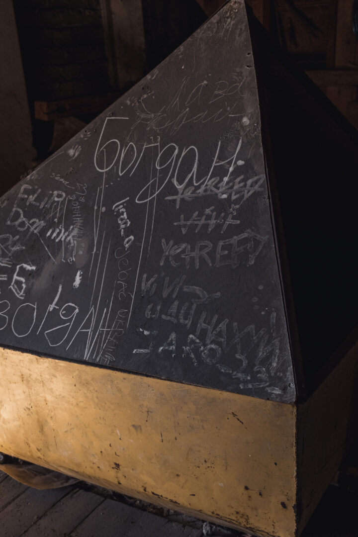Dark, triangular metal panel covered in chalk graffiti with random words and markings; yellow base at the bottom, in a dim workshop.