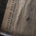 Old Polish newspaper page labeled 'MONITOR POLSKI' with dense columns of text and a decorative crest in the bottom-left corner, photographed at an angle.