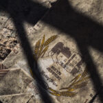 Dusty, torn paper showing a crest with wheat stalks and a building, lying on sandy ground.