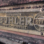 Close-up of a dirty, rusted metal nameplate on a vintage machine, with the large word 'BUTTER' embossed and worn edges nearby.