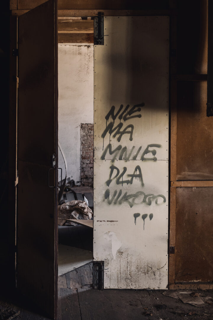 Dimly lit, abandoned doorway with a white metal door covered in black graffiti; piles of debris visible inside.